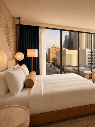 A stylish hotel room features a plush bed, modern lighting, and large windows showcasing a cityscape with tall buildings.
