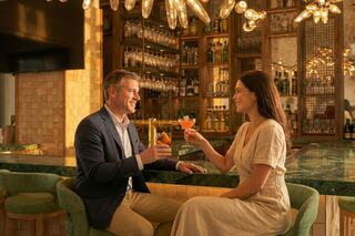 A couple enjoys drinks at a stylish bar, sharing smiles and laughter in a warm, inviting atmosphere. Elegant decor surrounds them.