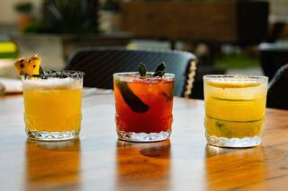 Three vibrant cocktails sit on a wooden table, showcasing yellow, red, and green drinks garnished with fruit and herbs against a sunny backdrop.
