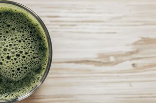 A frothy, green drink is seen in a clear glass, resting on a light wooden surface. The texture is smooth and vibrant.