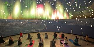A group practices yoga indoors, surrounded by a vibrant display mimicking the Northern Lights, creating a serene and immersive atmosphere.