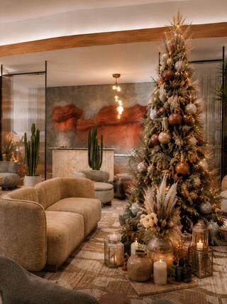 A beautifully decorated Christmas tree stands in a cozy, modern lounge, surrounded by plush seating and warm candlelight.