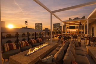 A chic rooftop lounge features cozy seating, an elegant fireplace, and a stunning sunset view over a city skyline.