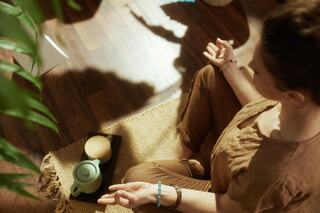 A person sits in a meditative pose on a woven mat, with a teapot and a candle nearby, surrounded by natural light and greenery.