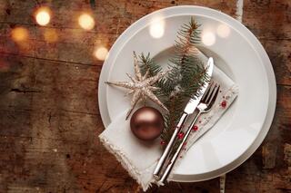 A decorative plate features a white napkin, silverware, a pine sprig, a holiday star, and an ornament, set against a rustic wooden background.