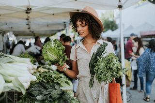 A woman in a hat examines fresh greens at a vibrant outdoor market, surrounded by fellow shoppers and colorful produce.