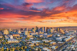 A stunning sunset casts vibrant colors over a sprawling city skyline, featuring modern buildings and a network of roads amidst greenery.