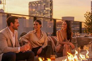 Three friends enjoy cocktails by a rooftop fire, overlooking a city skyline at sunset, sharing laughter and engaging conversation.