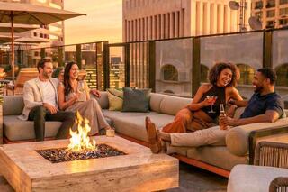 Four friends enjoy cocktails by a rooftop fire pit at sunset, surrounded by a stylish setting and city skyline. Laughter and camaraderie abound.