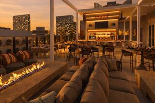 A stylish rooftop lounge features modern decor, cozy seating, a glowing fire pit, and a vibrant bar under a beautiful sunset sky.