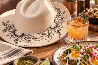A stylish hat adorned with floral designs sits beside a cocktail, dishes of vibrant food, and garnishes on a rustic wooden table.