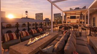 A stylish rooftop bar features cozy seating, a fire pit, and a vibrant city skyline at sunset, creating an inviting ambiance for relaxation.