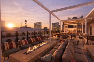 A chic rooftop lounge features cozy seating, a fire pit, and a vibrant city skyline at sunset, creating a warm, inviting atmosphere.