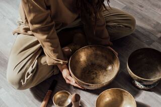 A person sits cross-legged, surrounded by various bowls and tools, with a focus on a large, shiny bowl held in their hands.