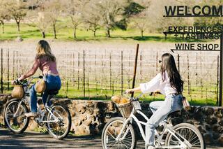 Two women ride bicycles past a vineyard, enjoying a sunny day, with a sign highlighting wine shop experiences and tastings.