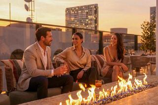 Three friends enjoy drinks on a rooftop at sunset, with a warm glow from a fire pit and a modern city skyline in the background.