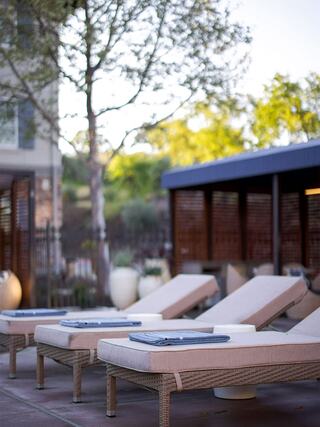 Lounge chairs with towels sit by a serene pool area, surrounded by greenery and a modern shaded structure, inviting relaxation.
