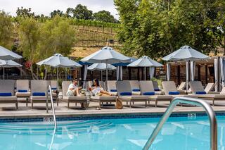 A couple relaxes by a poolside with lounge chairs and umbrellas, surrounded by greenery and vineyards in the background.