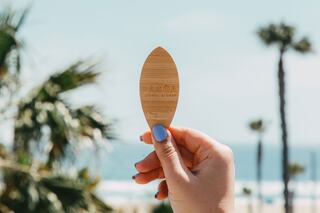 A hand holds a wooden paddle with the text "PACIFIC" against a sunny backdrop featuring palm trees and a beach view.