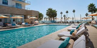 A sunny poolside scene with lounge chairs, umbrellas, and palm trees, overlooking a serene beach and clear blue water. Perfect for relaxation.