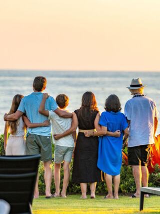 A group of seven people stands together at sunset, overlooking the ocean, embracing each other in a moment of togetherness.