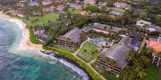 A tropical resort features lush greenery, palm trees, and a sandy beach, with pools and lounge chairs for relaxation by the ocean.
