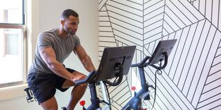 A man exercises on a stationary bike in a modern gym, focused and determined, with a stylish geometric wall design in the background.