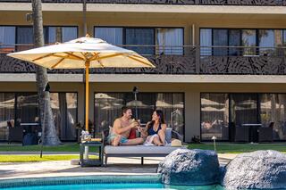 A couple enjoys drinks under an umbrella by the pool, lounging on a sunbed at a relaxing resort.