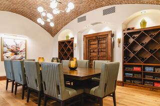 A stylish dining room features a wooden table surrounded by upholstered chairs, accented by warm lighting and wine storage on the walls.