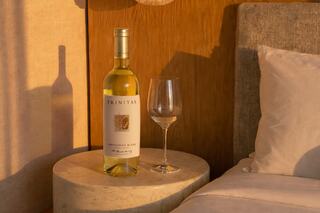 A bottle of white wine rests on a light-colored side table beside a glass and a cozy bed, illuminated by warm afternoon light.