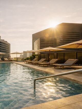 A tranquil poolside scene at sunset, featuring lounge chairs, sun umbrellas, and modern buildings reflecting warm golden light.