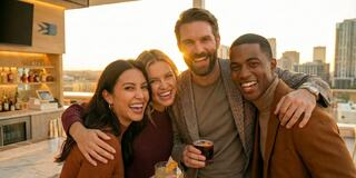 Four friends joyfully pose together at a rooftop bar during sunset, smiling and enjoying drinks against a city skyline backdrop.