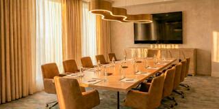 A modern conference room features a long wooden table, comfortable chairs, stylish lighting, and a large screen, surrounded by bright curtains.