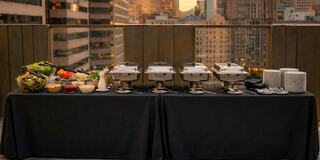 A buffet setup with salad, various dishes, and plates on a table, framed by a city skyline during sunset.