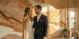 A happy couple shares a joyful moment, dressed elegantly, with warm sunlight illuminating the stylish interior behind them.