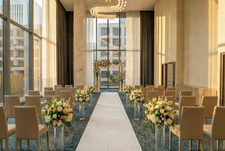 A beautifully decorated wedding venue with elegant floral arrangements, a white aisle, and soft lighting through large windows.