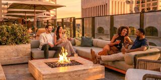 A group of four friends enjoy drinks and laughter around a fire pit on a stylish rooftop, with a sunset casting a warm glow.