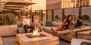 A group of four friends enjoy drinks and laughter around a fire pit on a stylish rooftop, with a sunset casting a warm glow.