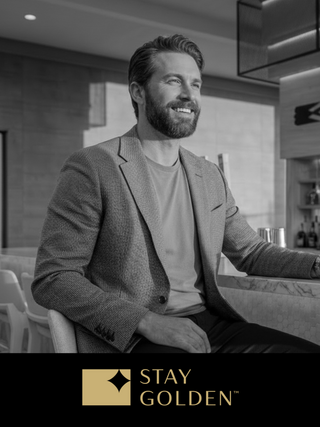 A smiling man in a stylish blazer sits at a modern bar, exuding confidence and charisma in a sophisticated, monochrome setting.