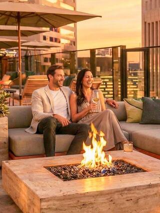 A couple enjoys drinks by a cozy outdoor fire pit, surrounded by stylish seating and a sunset view, radiating warmth and joy.