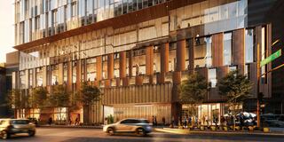 A modern building features large glass windows, wooden accents, and greenery in front, with cars and pedestrians on the street.