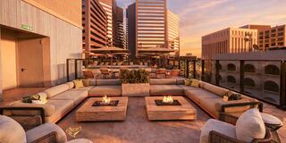 A stylish rooftop lounge features cozy seating around fire pits, surrounded by modern buildings at sunset, creating a warm, inviting atmosphere.
