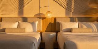 A stylish hotel room features two queen beds, soft lighting, and textured wall art, creating a warm and inviting atmosphere.