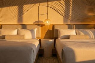 A serene bedroom features two elegant beds, cozy linens, modern decor, and warm golden sunlight streaming through the window.