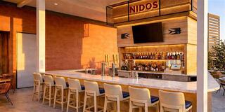 A stylish bar features a marble countertop, high stools, and a backlit shelf stocked with various beverages, all under warm lighting.