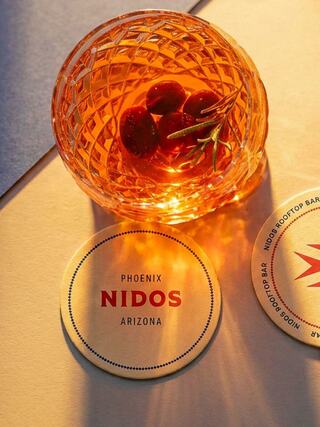 A crystal glass holds a amber drink garnished with olives and a sprig of rosemary, next to coasters featuring "Nidos" in Phoenix, Arizona.