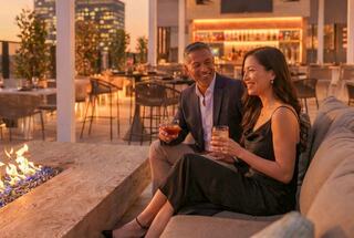 A couple enjoys cocktails by a cozy fire pit at a chic rooftop bar during sunset, sharing laughter and conversation.