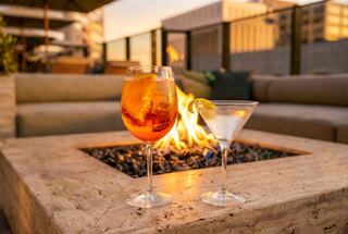 Two elegant cocktails sit beside a warm fire pit, with plush seating and a sunset glow creating a relaxing atmosphere.