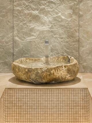 A sculptural stone basin with a smooth edge holds clear water, set against a textured wall and a tiled surface. A serene and natural atmosphere.