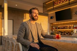 A smiling man sits at a bar during golden hour, enjoying a drink, radiating warmth and relaxation in a stylish setting.